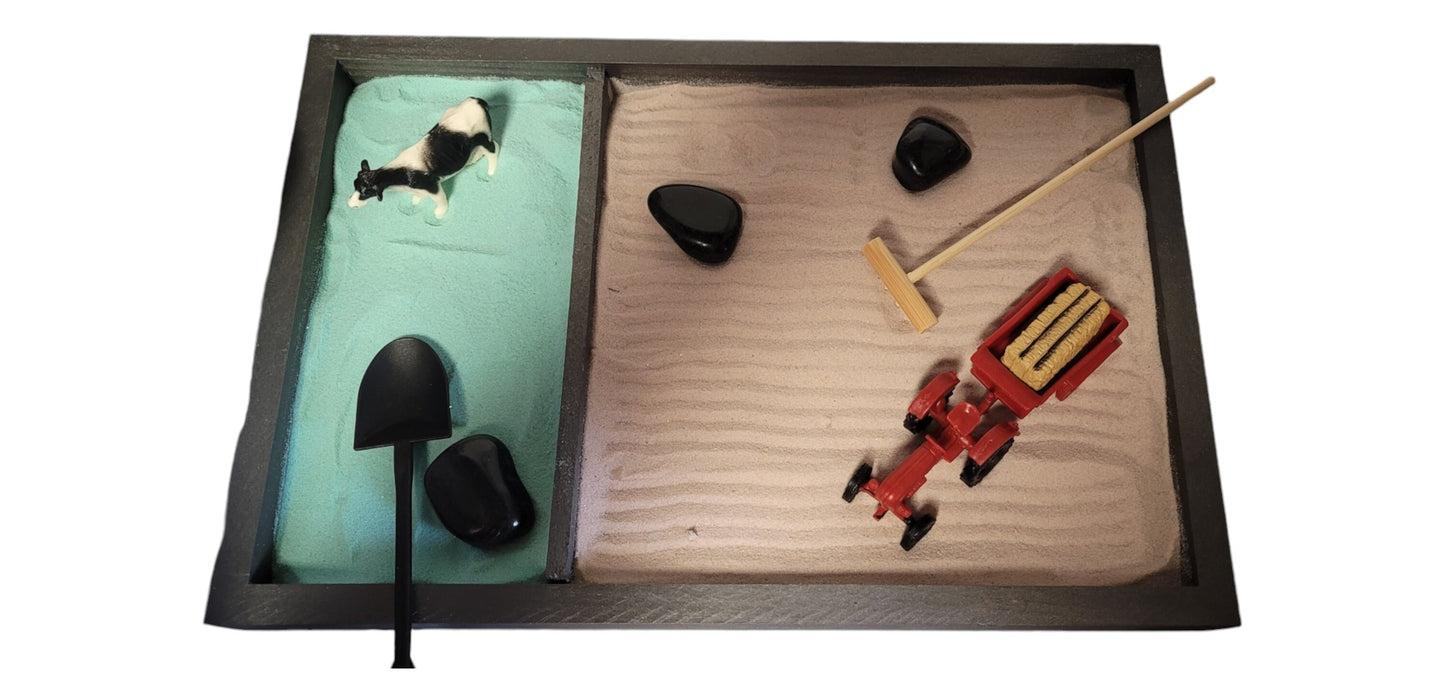 A Farm Zen Garden with a divided wooden tray, green and brown velvety sand, black rocks, a bamboo rake, and farm figurines.