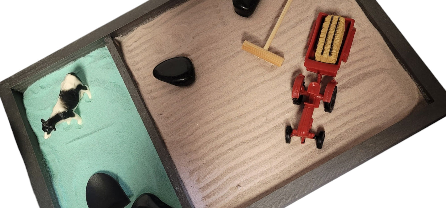 A Farm Zen Garden with a divided wooden tray, green and brown velvety sand, black rocks, a bamboo rake, and farm figurines.