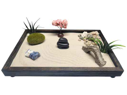 A traditional Zen garden set featuring a hand-crafted wooden tray with elegant feet, containing silky soft Zen sand, decorative elements including a bridge, tree, moss rocks, and grass bunches, along with a selection of polished black rocks, white marble stones, river pebbles, and a Sodalite healing crystal.