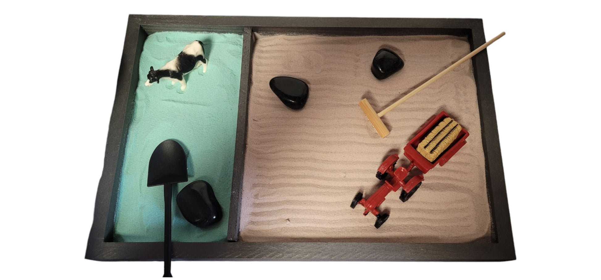 A Farm Zen Garden with a divided wooden tray, green and brown velvety sand, black rocks, a bamboo rake, and farm figurines.