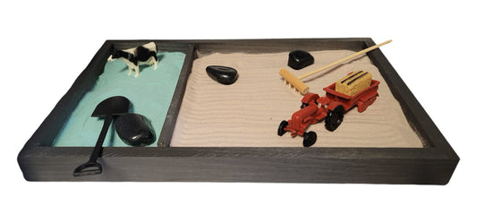 A Farm Zen Garden with a divided wooden tray, green and brown velvety sand, black rocks, a bamboo rake, and farm figurines.