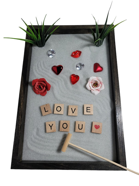 A Love Zen Garden with a bamboo rake, sparkling white sand, red and pink roses, hearts, and the words 'LOVE YOU' spelled out in letter tiles.