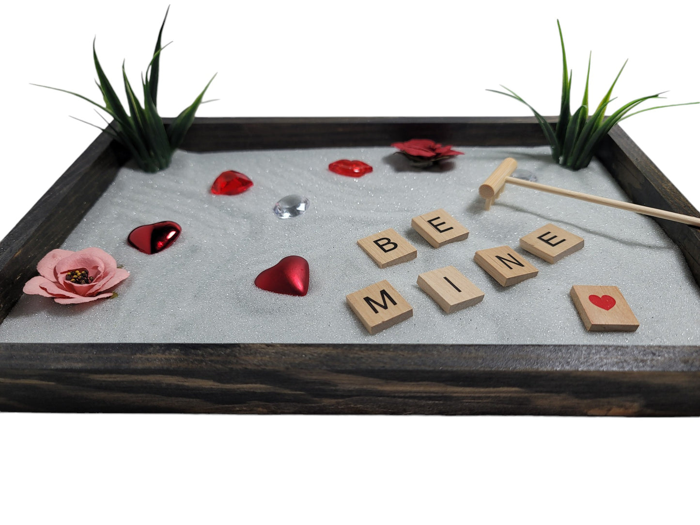 A Love Zen Garden with a bamboo rake, sparkling white sand, red and pink roses, hearts, and the words 'BE MINE' spelled out in letter tiles.