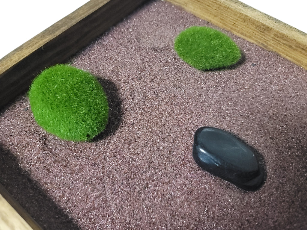 Zen garden with two green moss balls and one black polished stone on purple garnet sand.