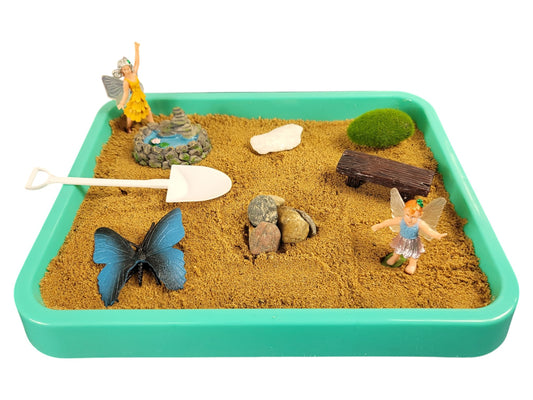 A Fairy Desktop Zen Garden with a teal tray containing sand, figurines of fairies and a butterfly, a wooden rake, and decorative elements like a pond, a bench, and stones.